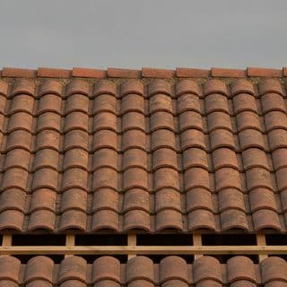 Een schuin dak met rode dakpannen, half gelegd op de panlatten zodat het houtwerk eronder zichtbaar is.