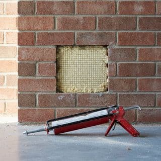 Een bakstenen gevel met een open plek waar de spouwisolatie zichtbaar is, en een kitspuit op de grond.
