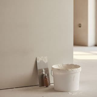 Een gladgestucte muur met een emmer stucmortel en gereedschap ervoor op de vloer.